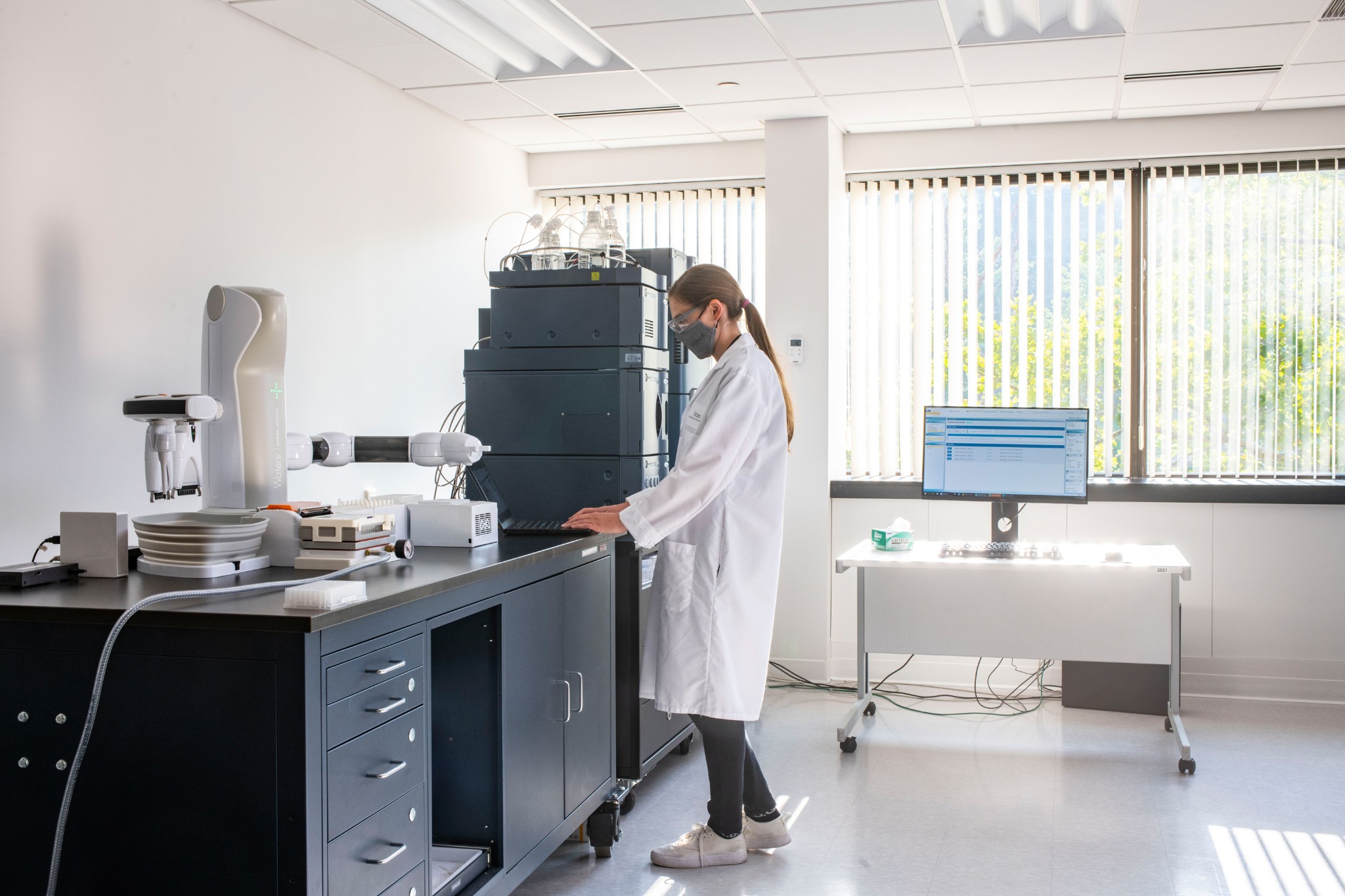 A scientist wearing a mask, standing at a laptop next to an Andrew+ Pipetting Robot