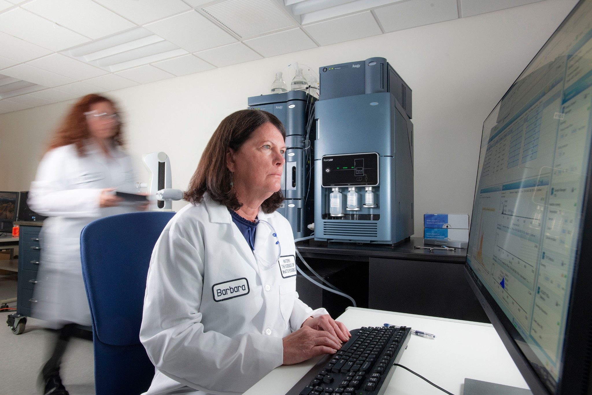 A scientist running a test on a computer in laboratory, with another scientist walking behind her