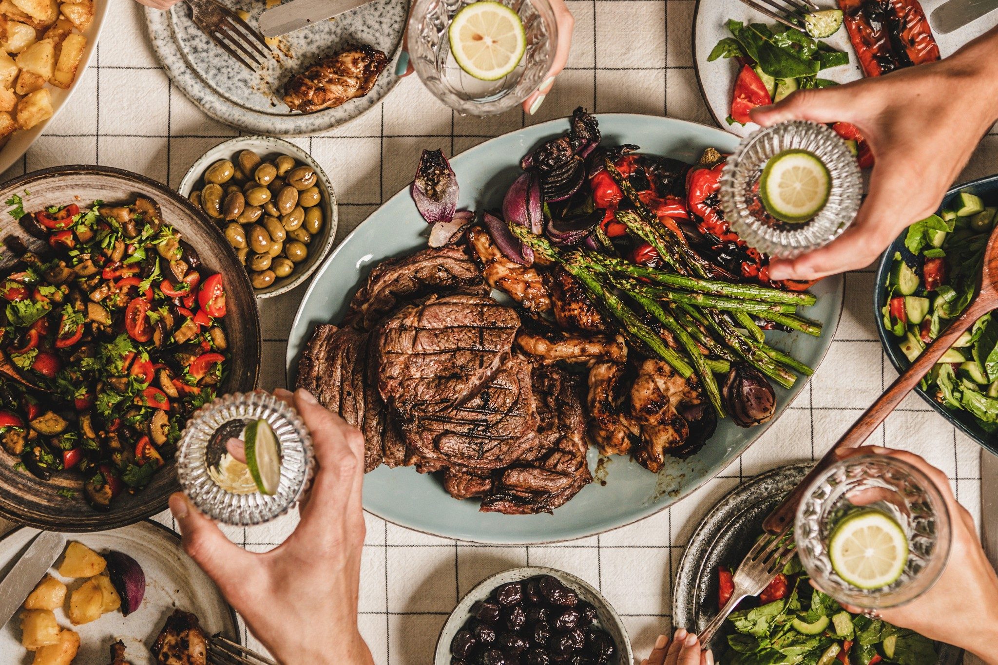 Summer barbeque party. Flat-lay of table with grilled meat, vegetables, salad, roasted potato and peoples hands with glasses of lemon water over white tablecloth, top view, wide composition