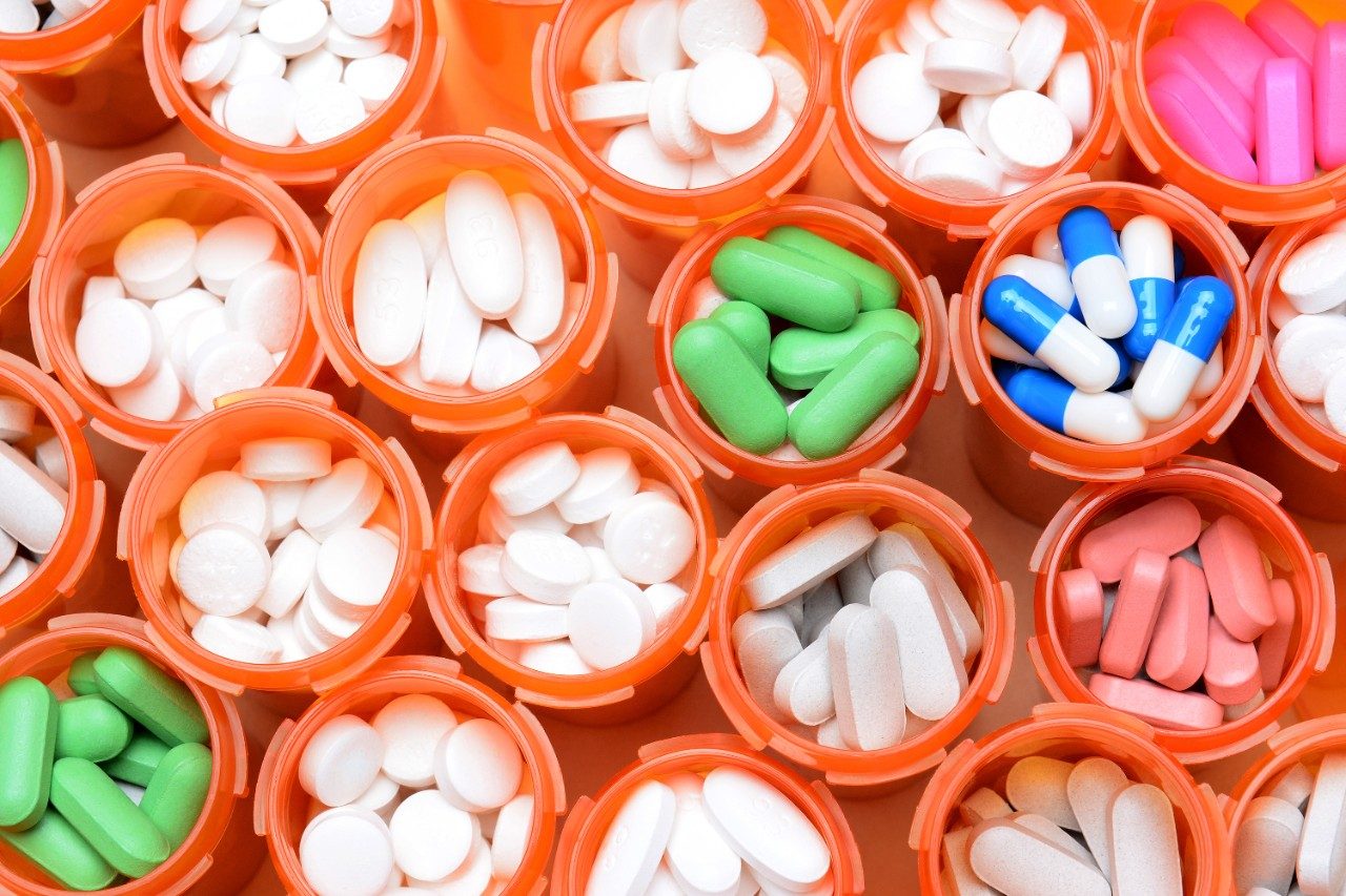 Looking down on a large group of prescription medicine bottles. The bottles all have their caps off and have a variety of drugs, tablets and capsules. Horizontal format filling the frame.