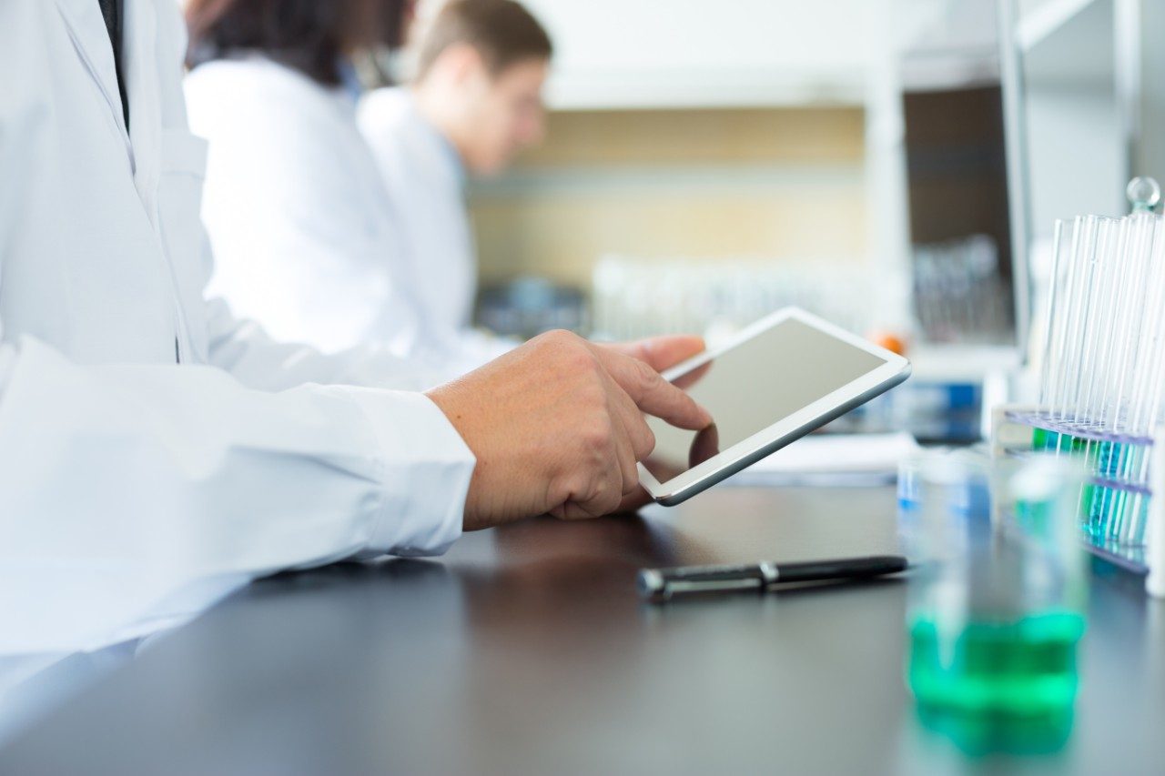 Portrait of Caucasian chemist in sterile uniform using tablet while standing next to machine with liquid soaps. Chemical factory interior.