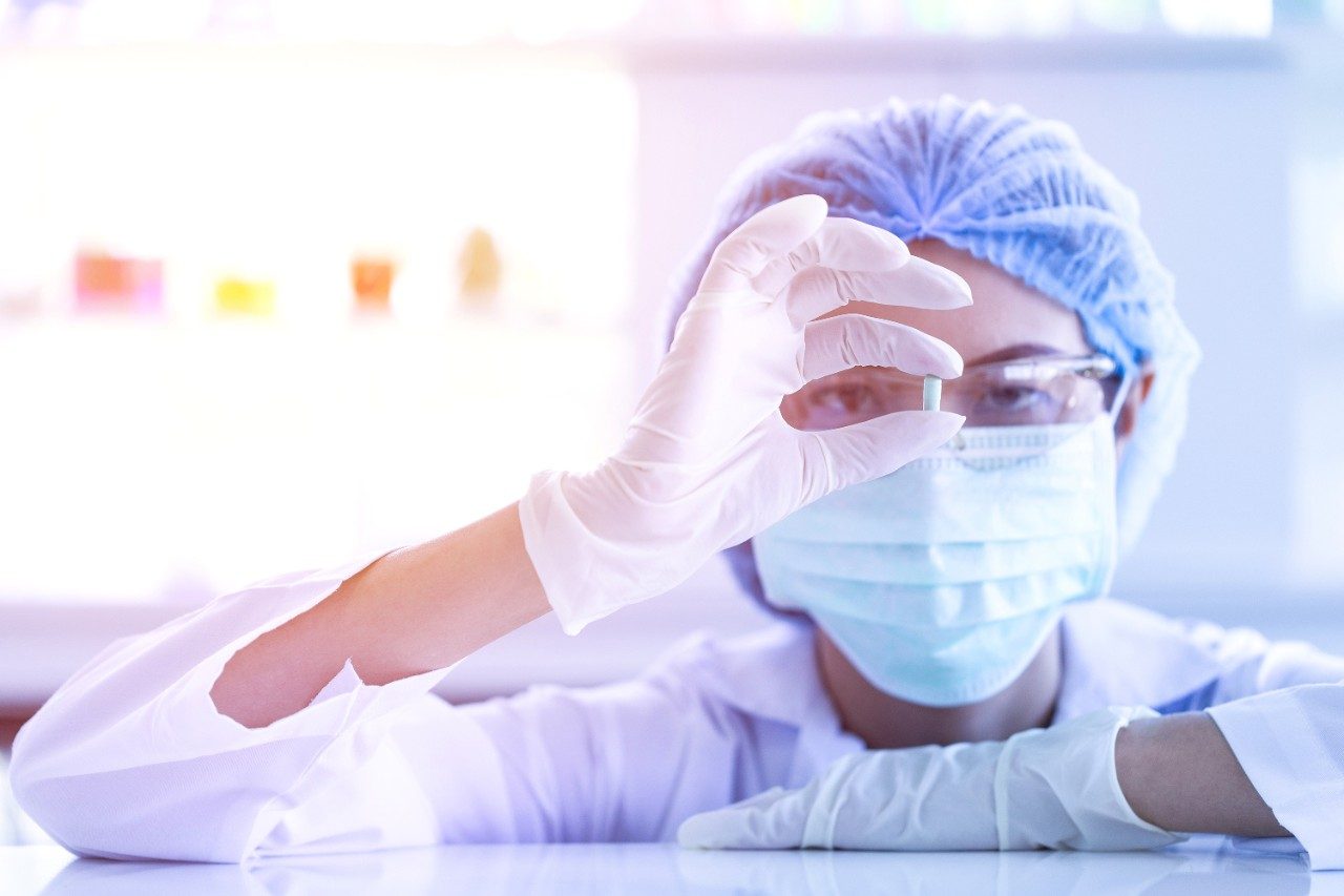 Close up shot of Asian female scientist holding small medicine capsule, there are blurred colorful test tubes, flask, colorful beakers be background.