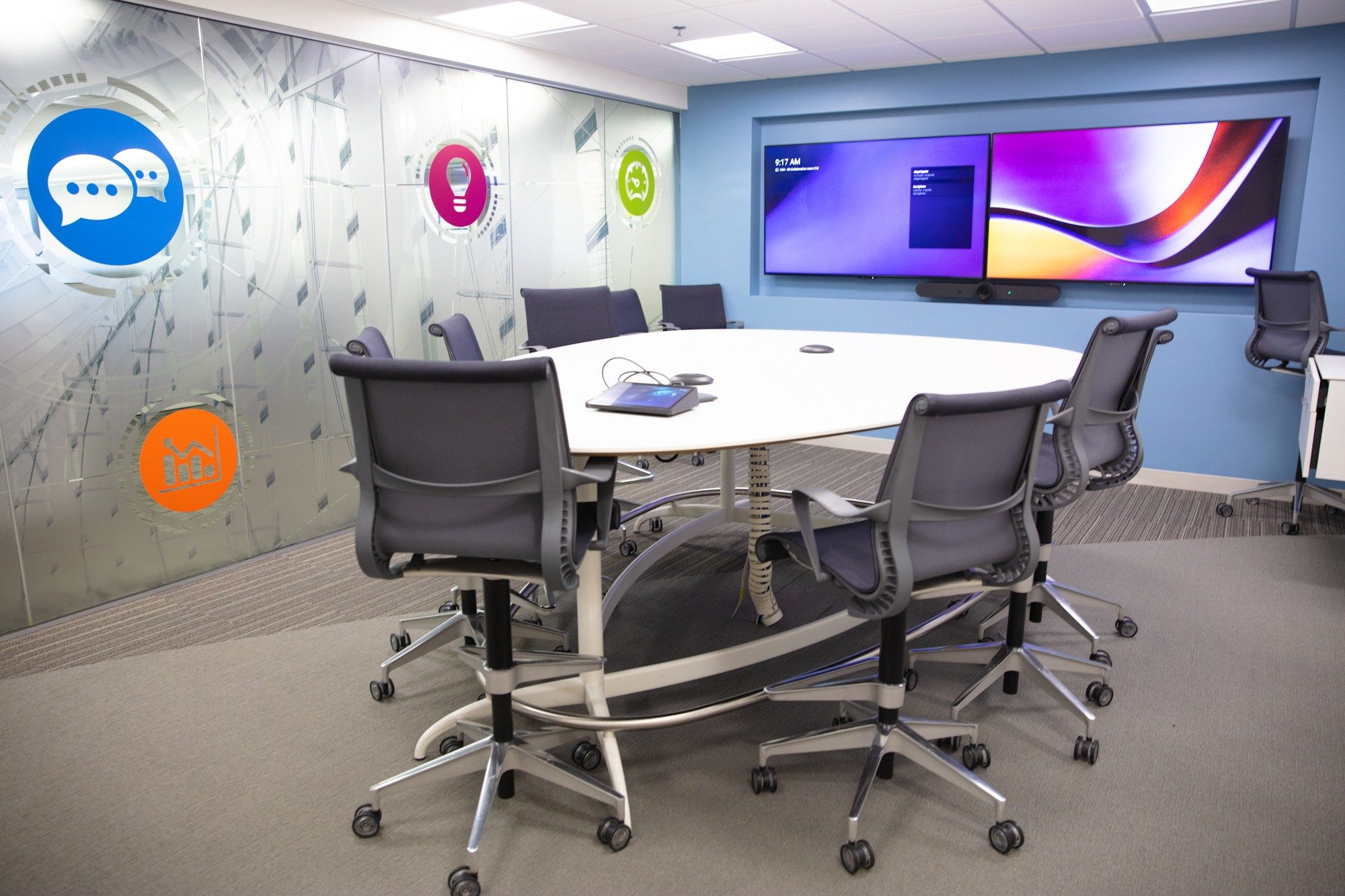 Sala de conferências, com uma grande mesa branca com várias cadeiras iguais e um par de monitores na parede.