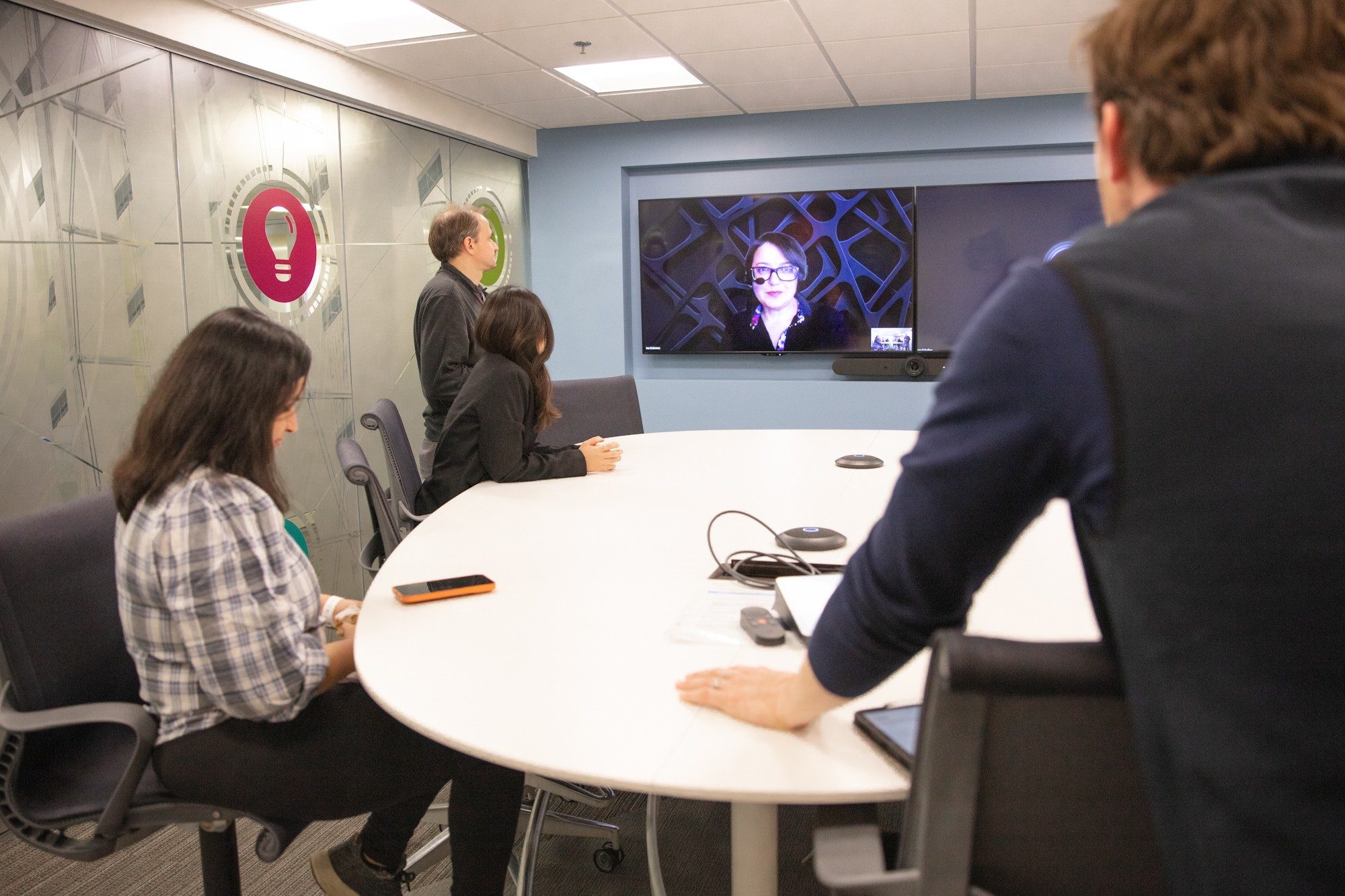 Quatro pesquisadores reunidos ao redor de uma mesa conversam com um participante virtual que aparece em um grande monitor.